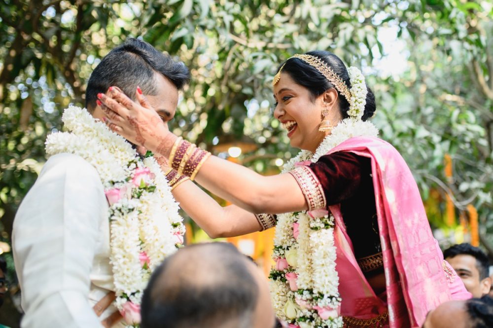 Bride playfully holding groom’s face during garland exchange at Tamil Brahmin wedding in Ecovillage, Pune